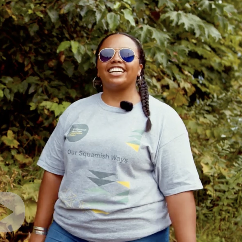 A smiling woman in sunglasses and a grey Our Squamish Ways T-shirt, standing among trees.